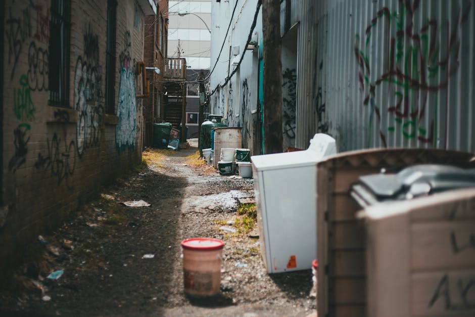 A middle-aged man with dark hair, wearing a black T-shirt with white text and dark trousers, is seen disposing of rubbish into a public metal refuse bin. He is bent over slightly, holding a large whit