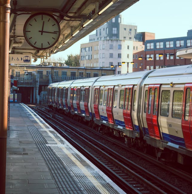 A moving train at an underground station platform, captured in a blurred motion shot highlighting the metallic exterior with a glossy finish. The train features a white lower section and a dark upper 